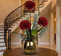 Élégante composition de chrysanthèmes rouges artificiels et d'eucalyptus dans un vase doré brillant, posée sur une table ronde en bois foncé dans un hall d'entrée luxueux avec un escalier à rampe en fer forgé et des colonnes en marbre.