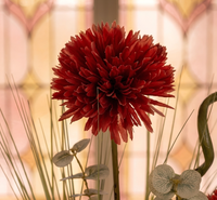 Gros plan sur une fleur de chrysanthème rouge bordeaux artificielle montrant la texture détaillée des pétales en tissu, accompagnée de feuilles d'eucalyptus givré et d'herbes fines, avec un arrière-plan flou aux tons chauds.