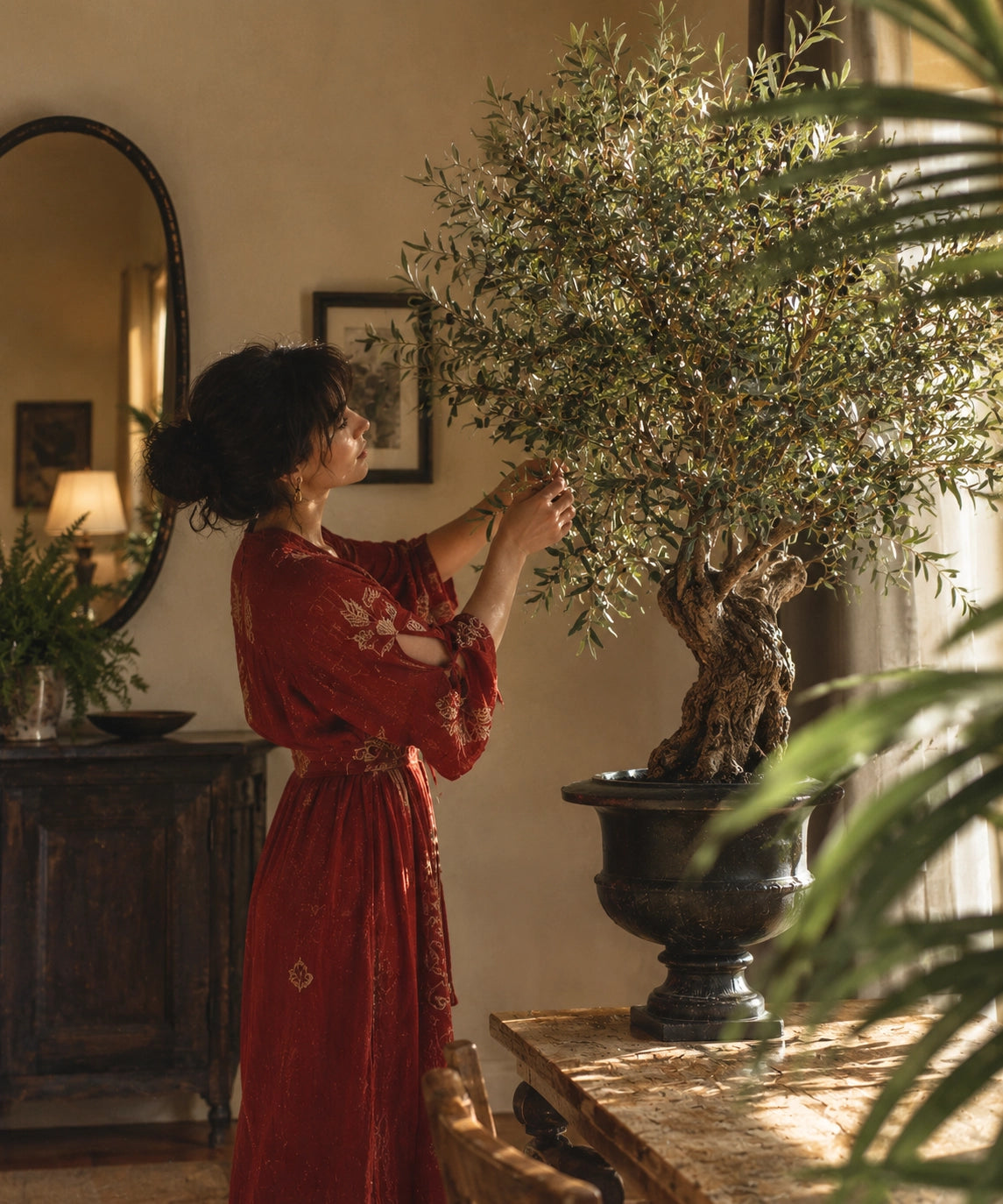 Une femme aux cheveux sombres attachés en chignon, vêtue d'une robe rouge à motifs, se tient à l'intérieur et s'occupe délicatement des feuilles d'un olivier artificiel luxuriant placé dans une urne sombre sur une table en bois, avec une lumière chaude du soleil traversant une fenêtre et un miroir ovale reflétant une lampe en arrière-plan.
