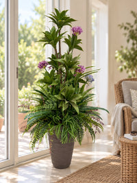 Composition florale et végétale artificielle avec agapanthes mauves dans un grand pot marron, posée près d'une baie vitrée lumineuse dans un salon moderne.