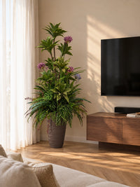 Grande composition florale artificielle avec agapanthes mauves et feuillages tropicaux, disposée dans un coin de salon ensoleillé près d'un meuble TV en bois et d'une fenêtre à rideaux voilages.