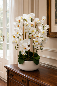 Composition florale artificielle d’orchidées blanches dans un pot blanc décoratif, exposée sur une console en bois dans un salon élégant au Maroc – Sara Flora