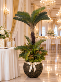 Palmier cycas artificiel effet réel dans un pot doré orné d'un nœud en ruban crème, décoration d'une salle de réception de mariage de luxe avec lustres et tables dressées