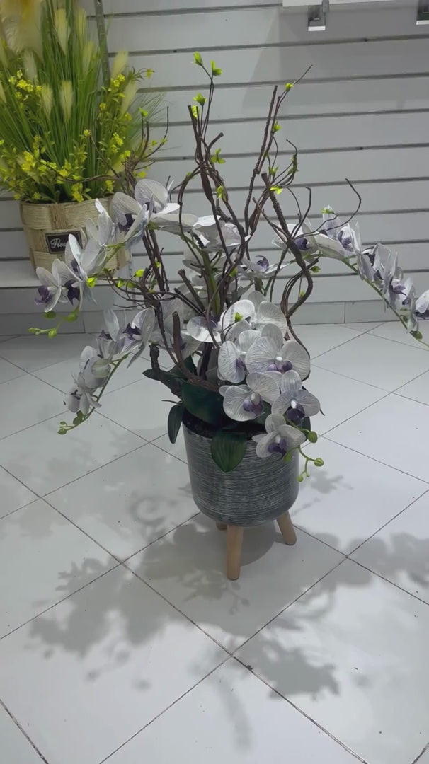 Orchidées blanches et violettes artificielles dans leur pot décoratif sur pieds en bois, présentées dans le showroom SaraFlora au Maroc